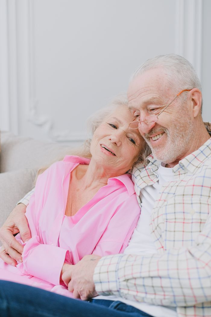 Elderly couple sitting together, smiling warmly and displaying affection indoors.